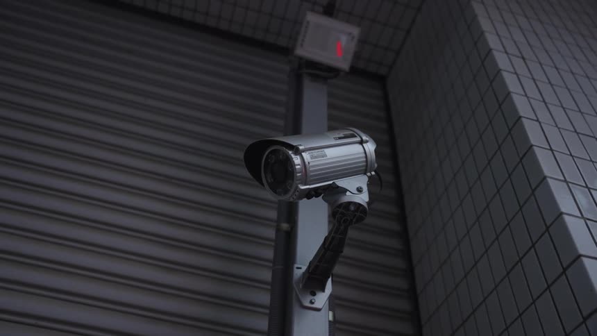 A low-angle shot of a CCTV security camera with a blinking red light mounted on a building at night