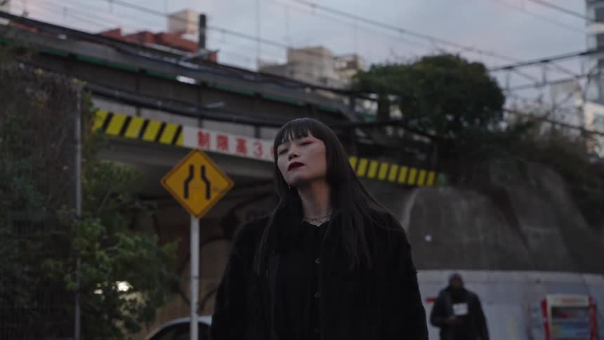 A stylish Japanese woman in a black coat closes her eyes before looking directly at the camera on a moody urban street