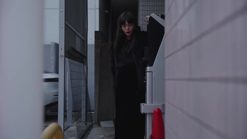 A chic Japanese woman in a black fur coat strikes a dramatic pose against a payment kiosk in a moody city alley