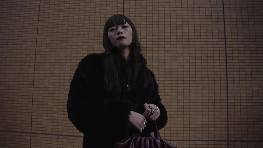 A slow zoom on a stylish Japanese woman in a black fur coat posing confidently against a tiled urban wall
