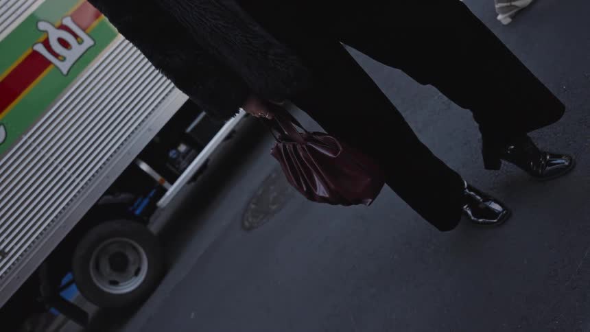 A slow motion, low-angle shot of a stylish woman in a black fur coat holding a burgundy handbag while waiting at a city crosswalk