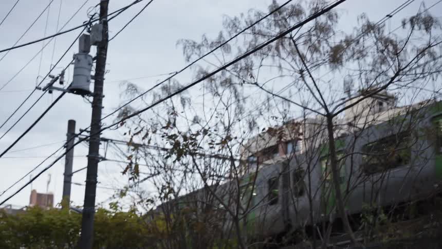 A panning shot follows a green and silver commuter train speeding through an urban area in Japan