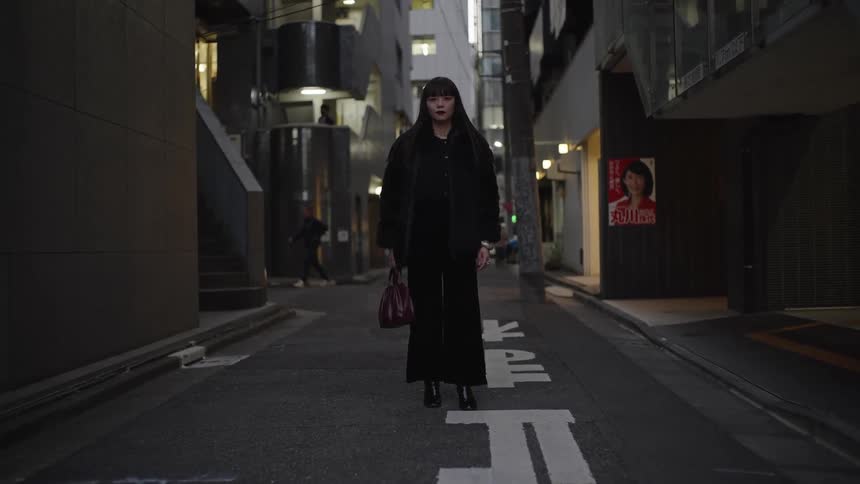 A stylish woman in a black outfit stands still in the middle of a dark Tokyo alleyway at night