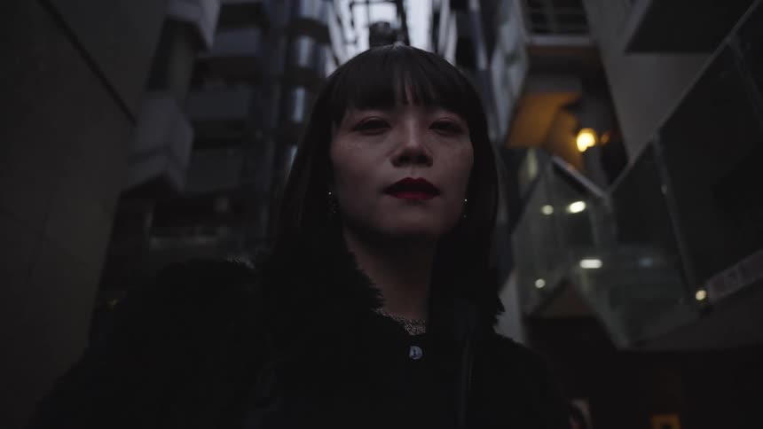 Low angle slow-motion shot of a stylish Asian woman looking at the camera while standing in a dark, modern city alley