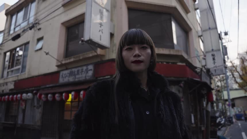 A slow zoom on a stylish Japanese woman in a black fur coat standing on a city street corner and staring intensely at the camera