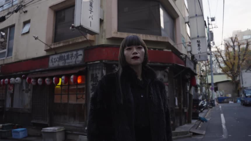 A fashionable Japanese woman in a black fur coat stares into the camera while standing on a quiet street corner in Tokyo