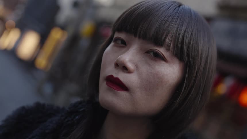 A slow-motion, close-up portrait of a stylish Asian woman with a bob haircut looking seriously on a city street at dusk
