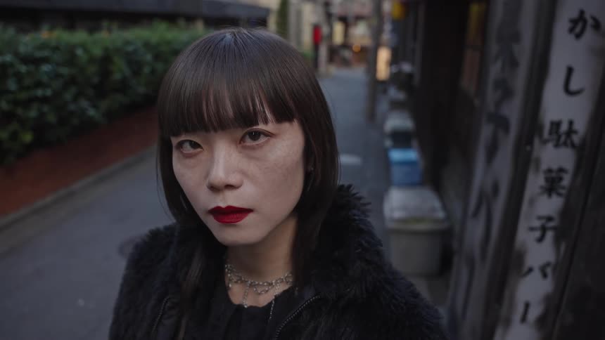 Close-up portrait of an edgy Asian woman with bangs and red lipstick staring intently into the camera in a narrow Tokyo alleyway