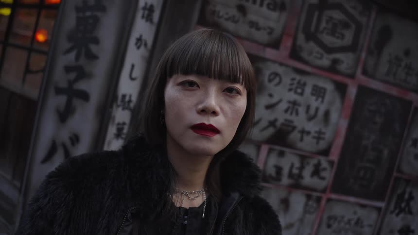 A stylish Japanese woman with bold red lipstick and a black coat stares intensely at the camera in a slow-motion portrait on a city street