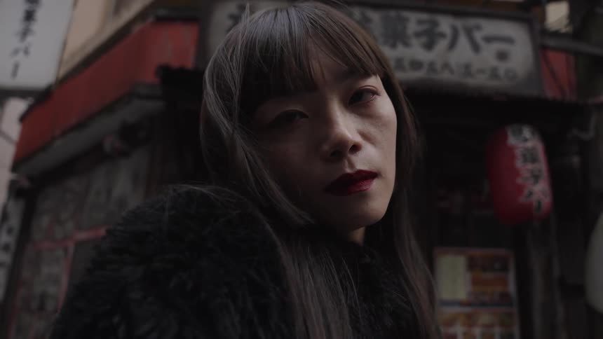 A mysterious Japanese woman with dark lipstick stares intensely as the wind blows through her hair on a moody city street in slow motion