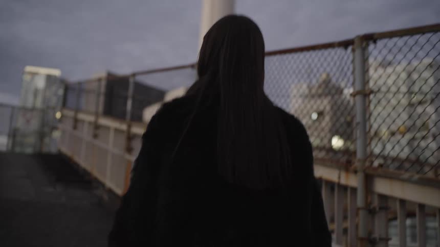 A stylish Asian woman in a black fur coat walks along an urban walkway at dusk in slow motion