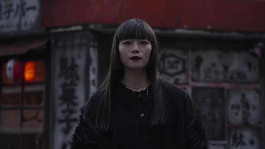 A stylish Asian woman with dark bangs and red lipstick slowly closes and opens her eyes on a moody city street in Japan