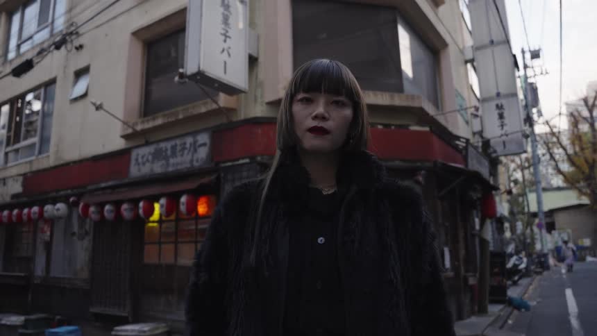A stylish Japanese woman in a black fur coat stares into the camera on a Tokyo street corner as it slowly pushes in