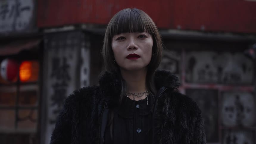 An intense slow zoom portrait of a stylish Japanese woman in a black fur coat staring directly into the camera on a moody city street