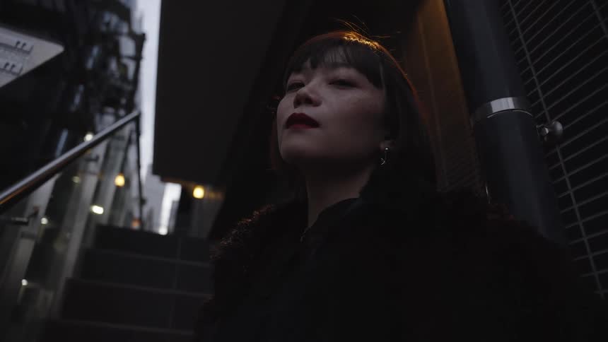 A mysterious and stylish young Asian woman sits pensively under a light on dark city stairs at night