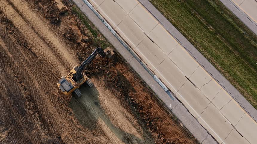 A stationary top-down aerial view shows an excavator digging a trench next to a busy highway with blurred traffic speeding by