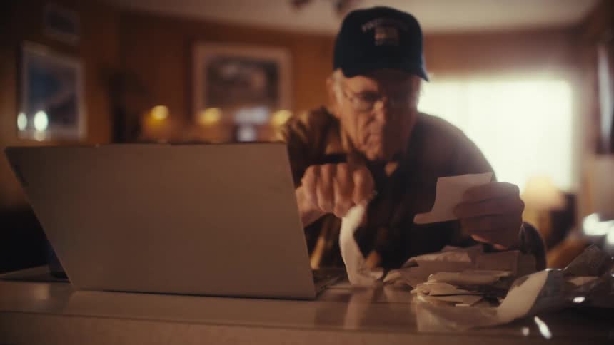 An elderly man in a hat carefully organizes his receipts and bills while working on his finances with a laptop at home