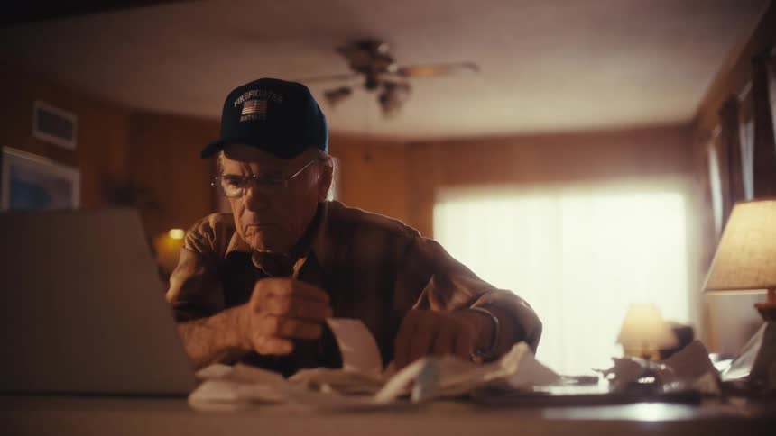 Focused elderly man wearing a retired firefighter hat meticulously sorts through a pile of receipts while working on his finances at a laptop
