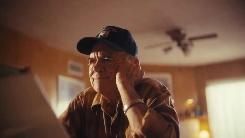 A happy elderly veteran smiles as he uses a laptop in his cozy, sunlit home
