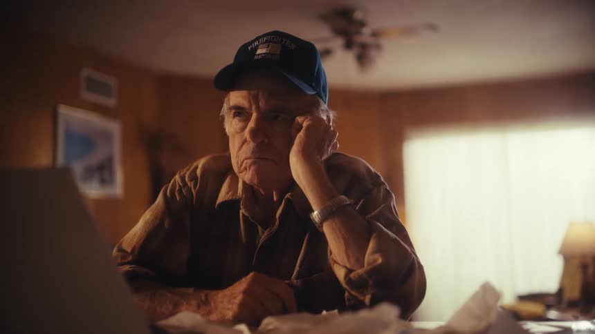 A stressed elderly retired firefighter sits at his desk overwhelmed by bills and financial worries while working on his laptop