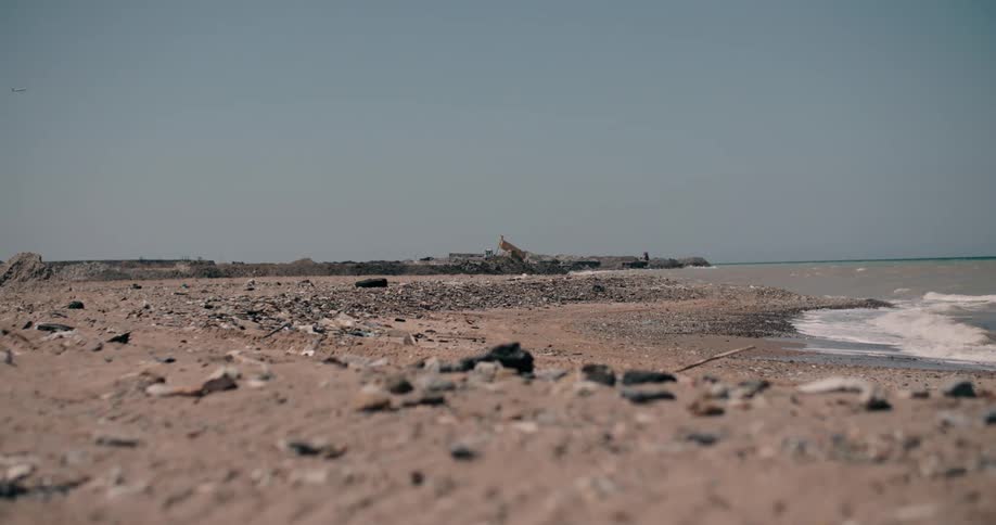 A low-angle, static shot captures a polluted, rocky beach with gentle ocean waves as an excavator works in the distance