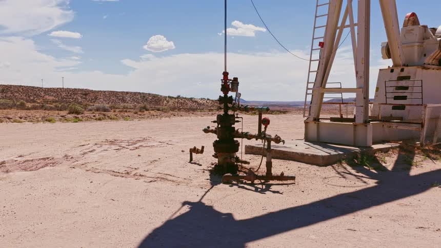 An oil pump jack operates in a vast, arid desert landscape under a bright blue sky