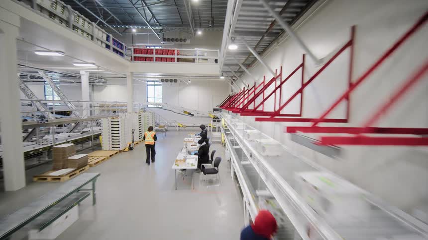 A high-angle tracking shot follows a bustling automated conveyor belt system in a modern Canadian distribution warehouse