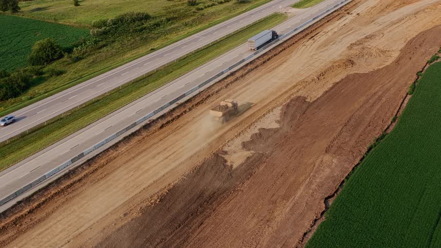 Aerial drone view of a large dump truck moving earth along a dusty construction site next to a busy highway in the countryside
