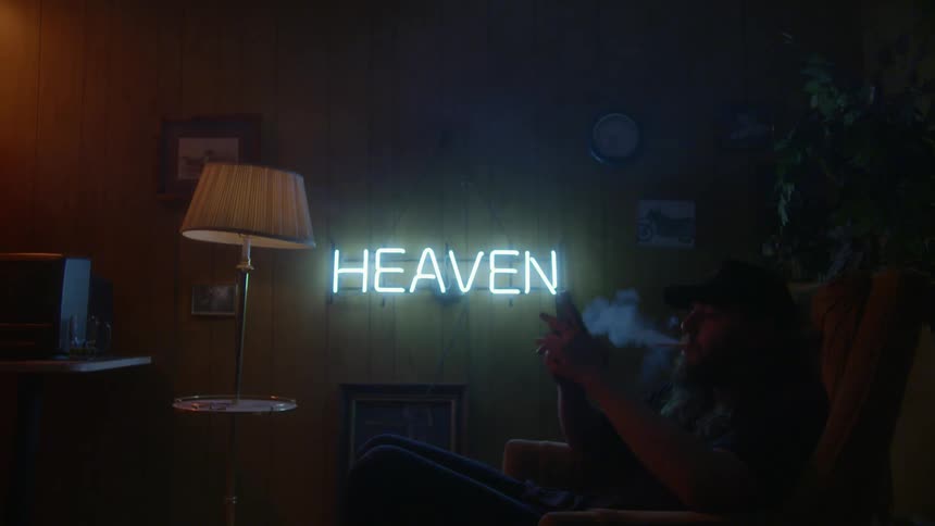 Man sits smoking on a couch in a dim room lit with a neon sign