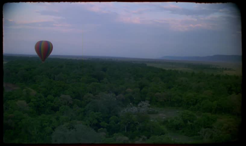 Vintage aerial footage of a single colorful hot air balloon drifting over a vast, dense green forest at dawn