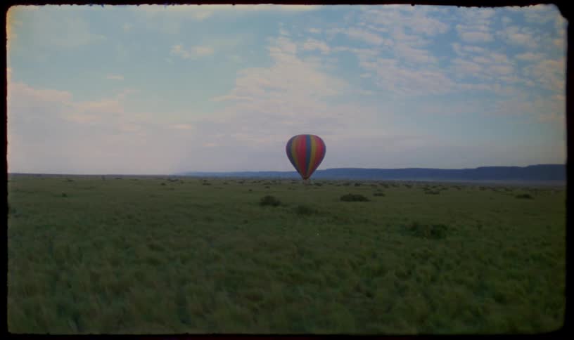 A vintage aerial view shows a colorful hot air balloon drifting peacefully over a vast, green savanna