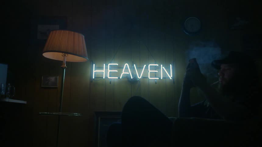 Man sits on a couch in dim room with neon sign