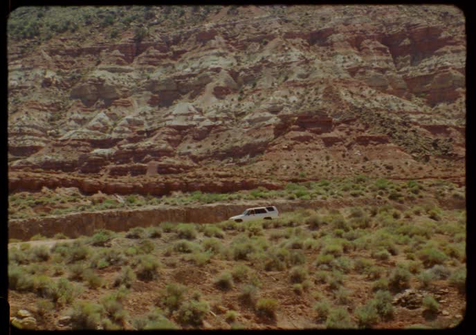 Vintage Super 8 footage shows a white SUV driving on a dirt road through the vast, arid landscape of a red rock canyon