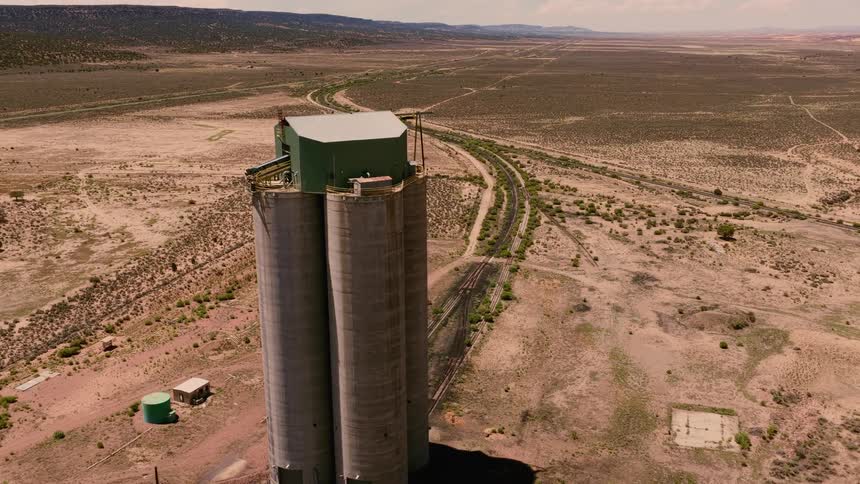 An aerial drone shot flies past a large industrial silo and railroad tracks in the vast, arid desert landscape