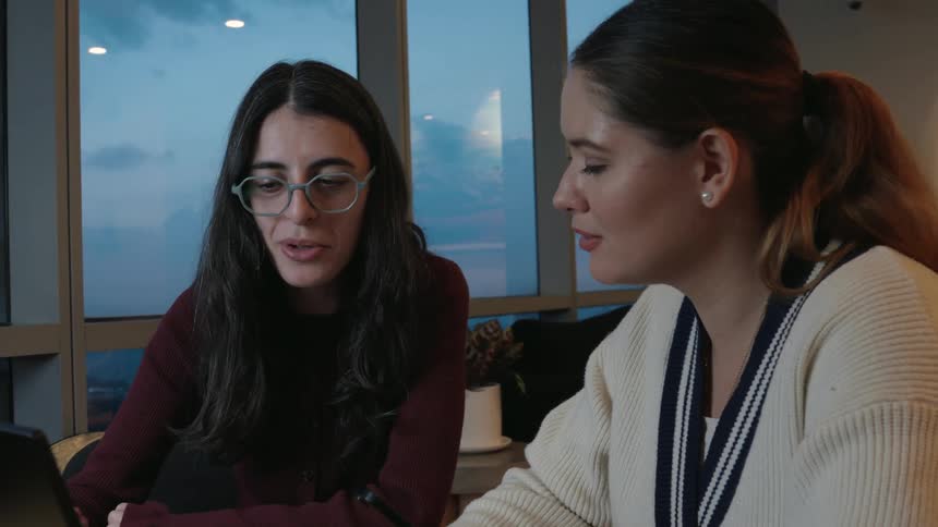 Two female colleagues are deep in conversation while working together on a laptop in a modern office with a city view at dusk