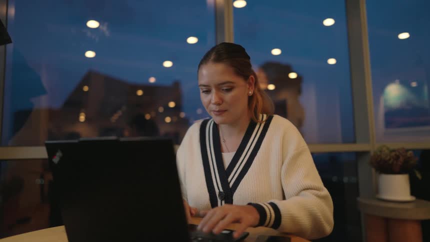 A young businesswoman smiles while working on her laptop in a modern office space in the evening