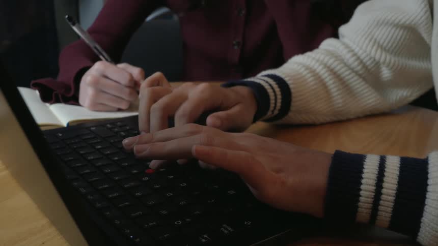 A close-up of one person's hands typing on a laptop keyboard while another person takes notes in a notebook beside them