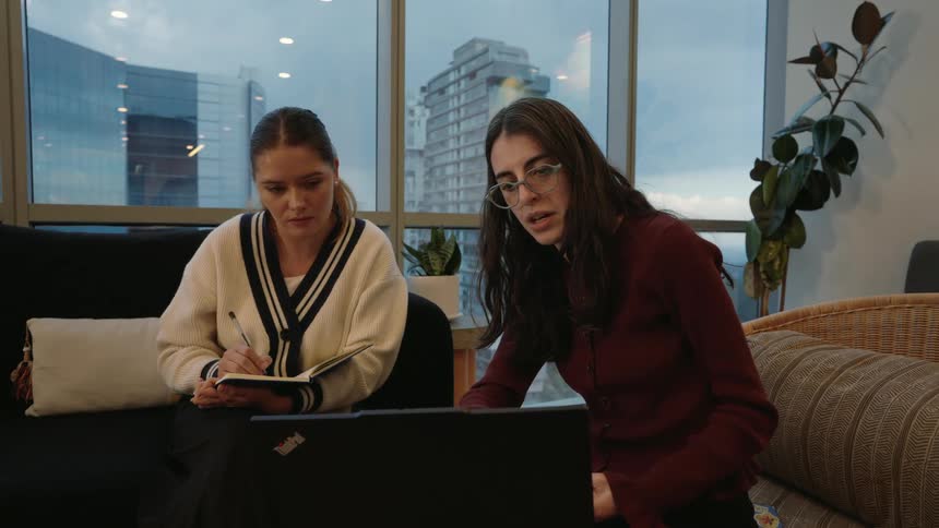 Two young female colleagues collaborate on a project, discussing details on a laptop while one takes notes in a modern office lounge