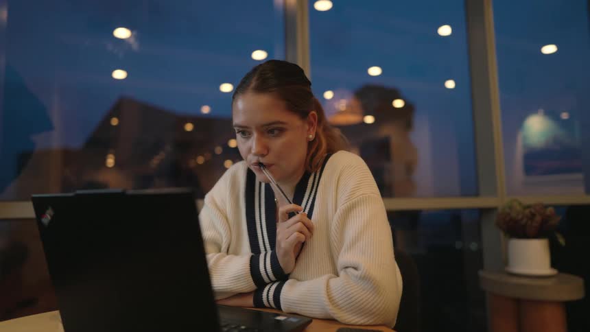 A focused young woman works late on her laptop, thoughtfully holding a pen to her mouth as she contemplates her work