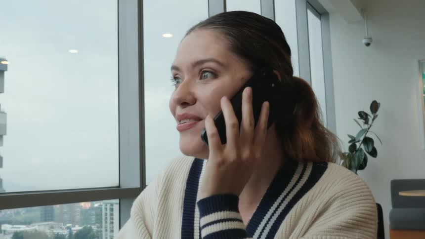A beautiful young woman working in a modern office with a city view smiles as she receives an exciting phone call