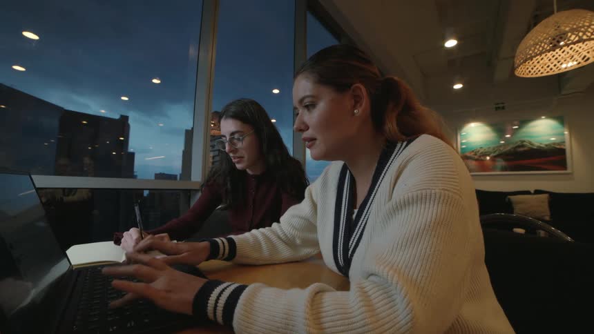 Two young women collaborate on a project in a modern office, working on a laptop with a city view at dusk