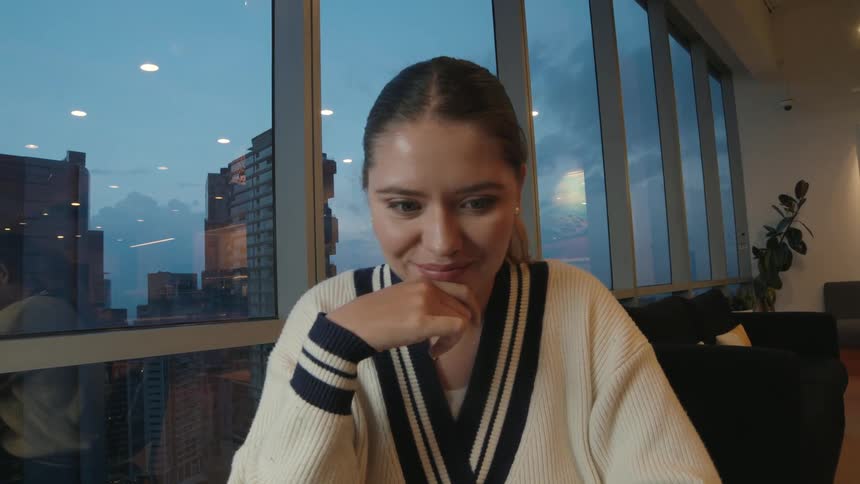 A thoughtful young woman works on her laptop in a modern office overlooking the city at dusk