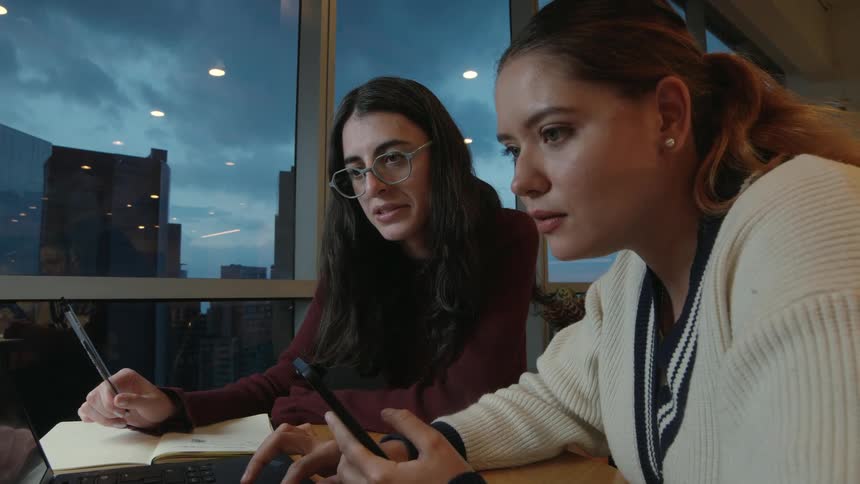 Two young women collaborate on a project using a laptop in a modern office with a city view at dusk