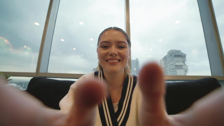 POV shot of a happy young woman smiling and reaching for the camera while sitting in a modern office