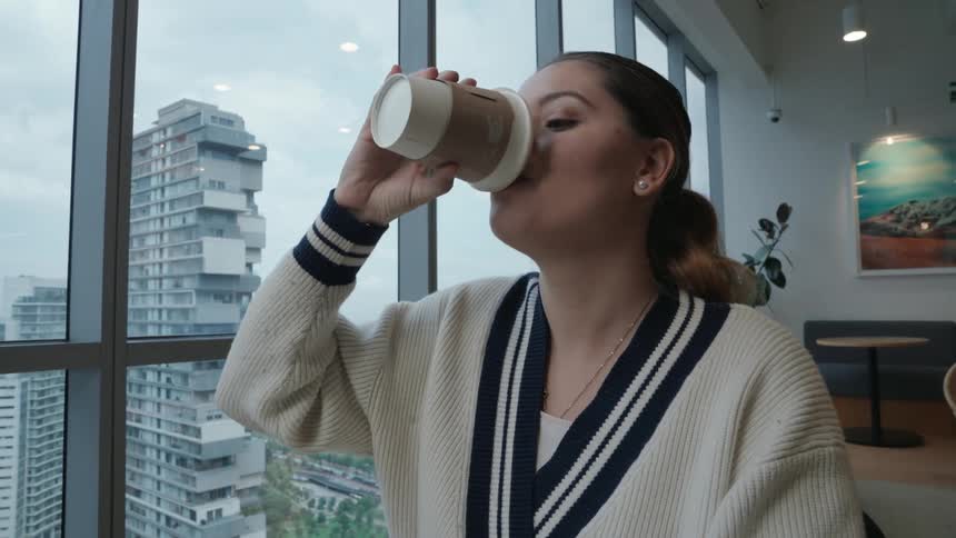 A young professional woman smiles while working on her laptop and taking a coffee break in a modern high-rise office with a city view