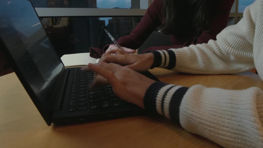 A person's hands type on a laptop keyboard while another person writes in a notebook beside them