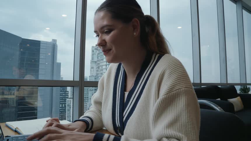 A smiling young woman takes notes and works on her laptop at a desk in a modern high-rise office with a city view