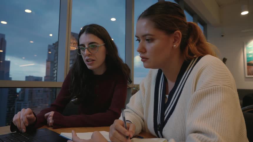 Two young women work together on a laptop in a modern office with a city view at twilight