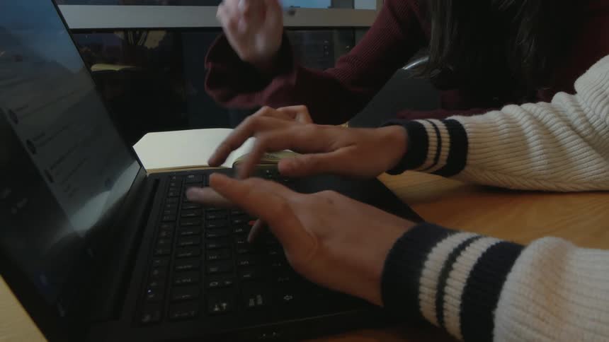 Close-up of two people working together, one typing on a laptop keyboard while the other points at the screen with a pen