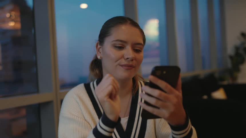 A pleasant young woman leisurely scrolls and taps on her smartphone in a modern room with a city view at twilight